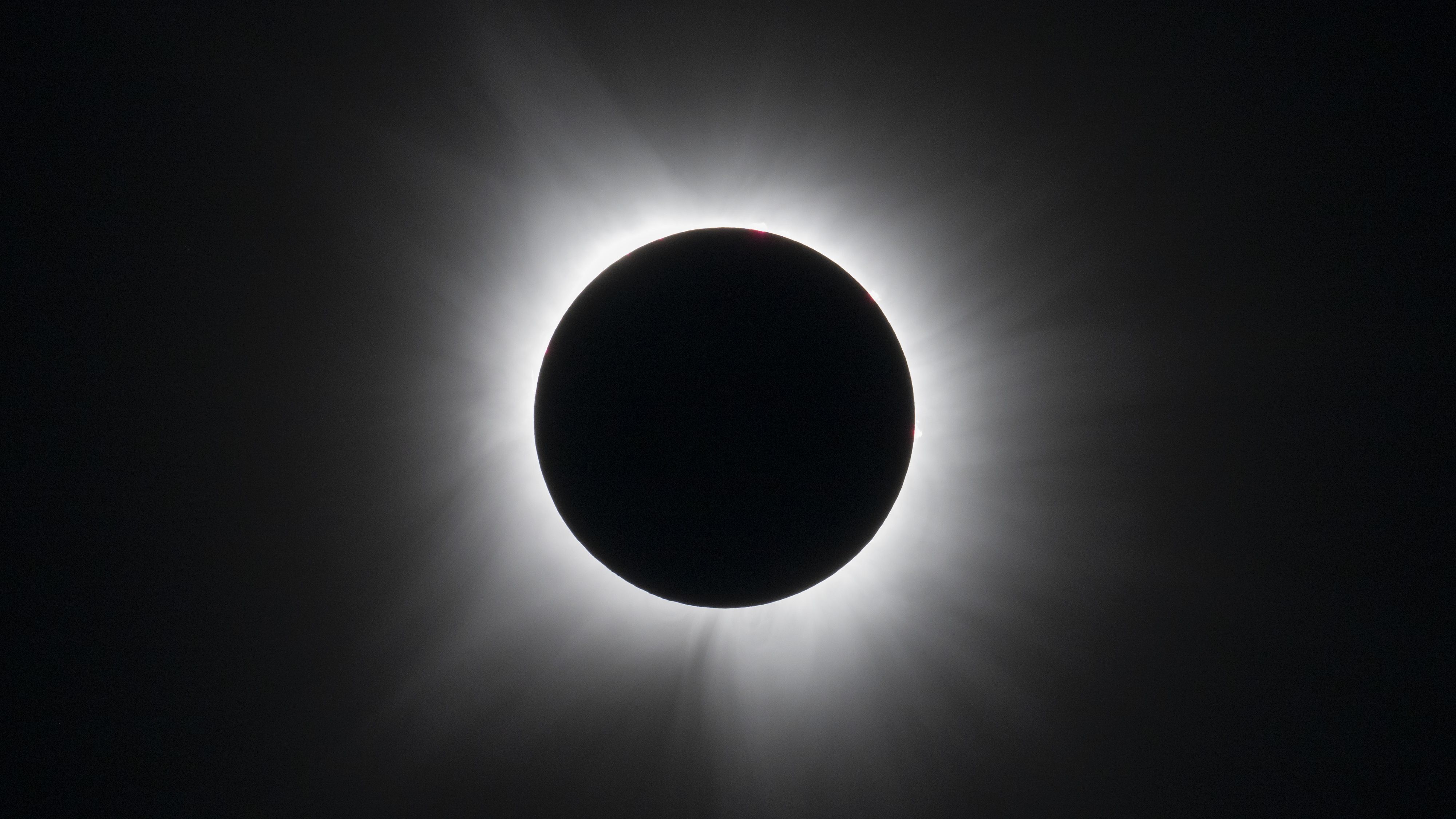The moon is photographed blocking the sun's disk during totality at the climax of a total solar eclipse. The sun's white atmosphere haloes the moon in a black sky.