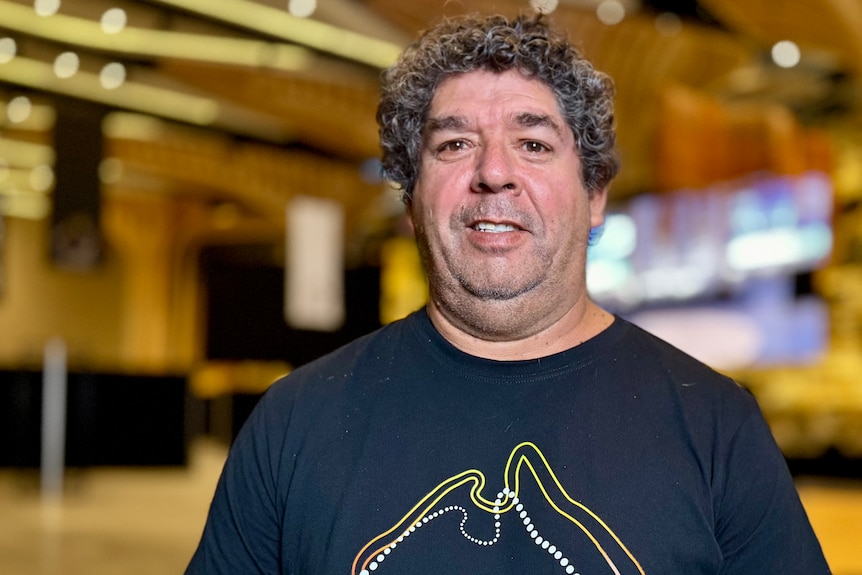 A man wearing a black t-shirt with an outline of Australia and the words 'National Indigenous Art Fair' stands in a large room.