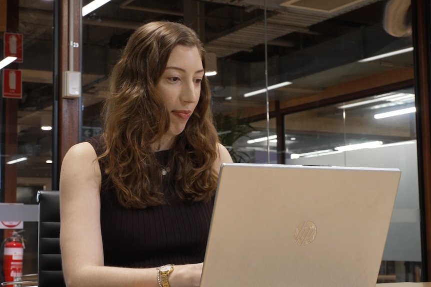 A dark-haired woman in a dark, sleeveless top operates a laptop while sitting at a desk in an office.