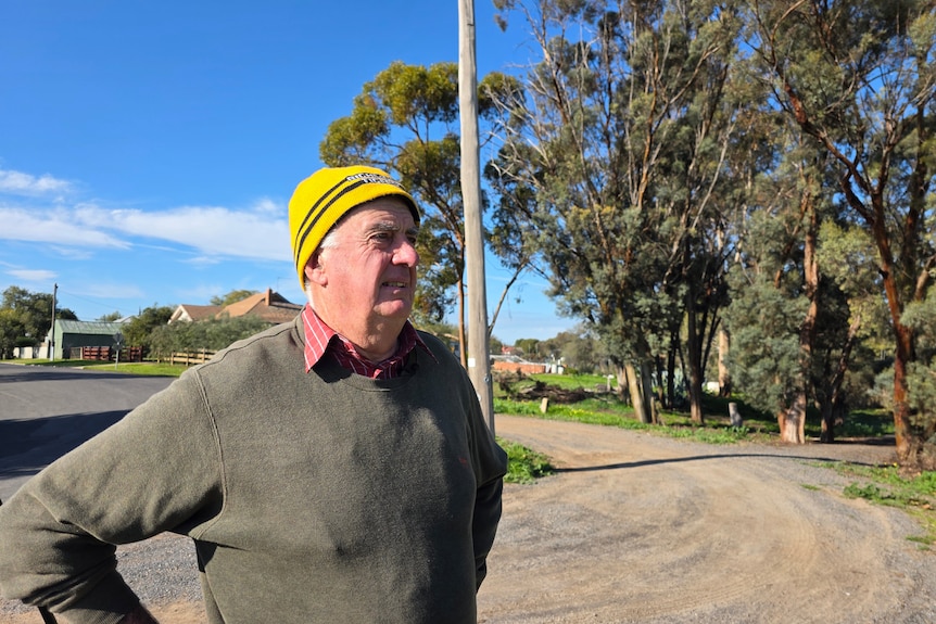 A middle-aged man in a beanie and a jumper stands on the side of a suburban street.