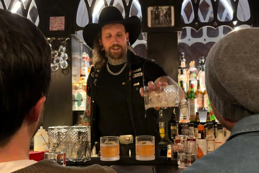 Man in dark bar, clothes and hat pouring drink into glasses between two people