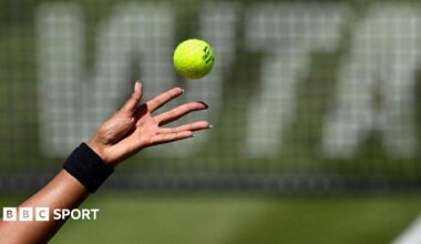 A female tennis player with long fingernails (only the hand shown) tosses a tennis ball in front of a net featuring WTA