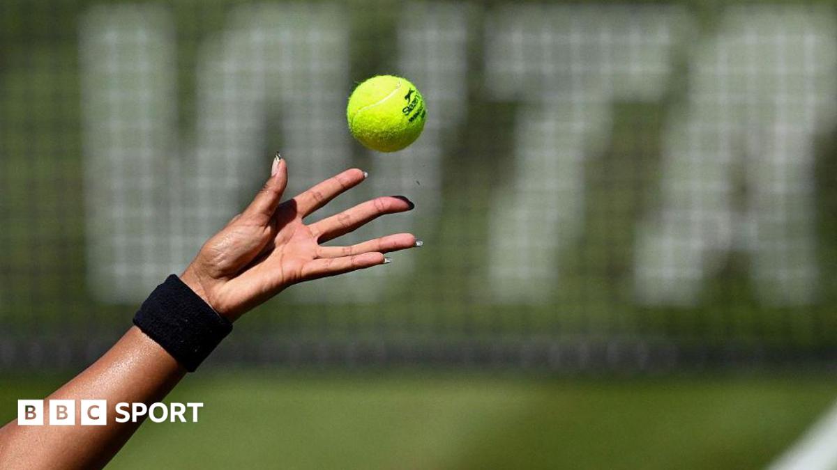 A female tennis player with long fingernails (only the hand shown) tosses a tennis ball in front of a net featuring WTA
