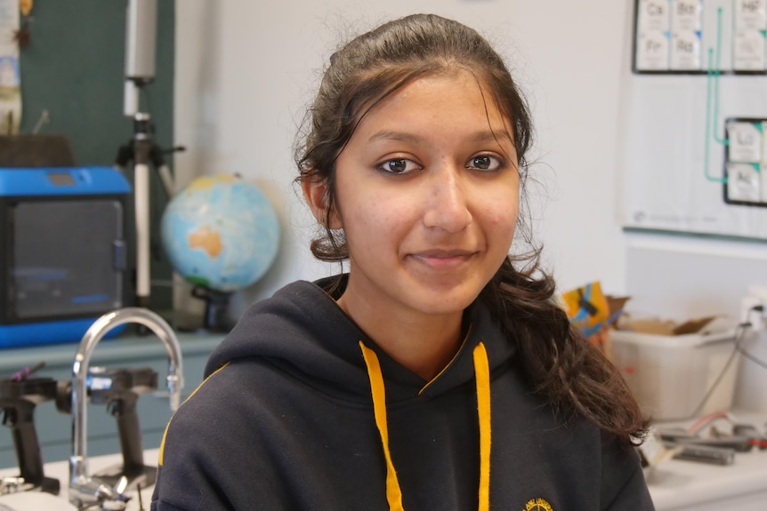 A girl with dark skin sitting in a science lab with a school jumper on.