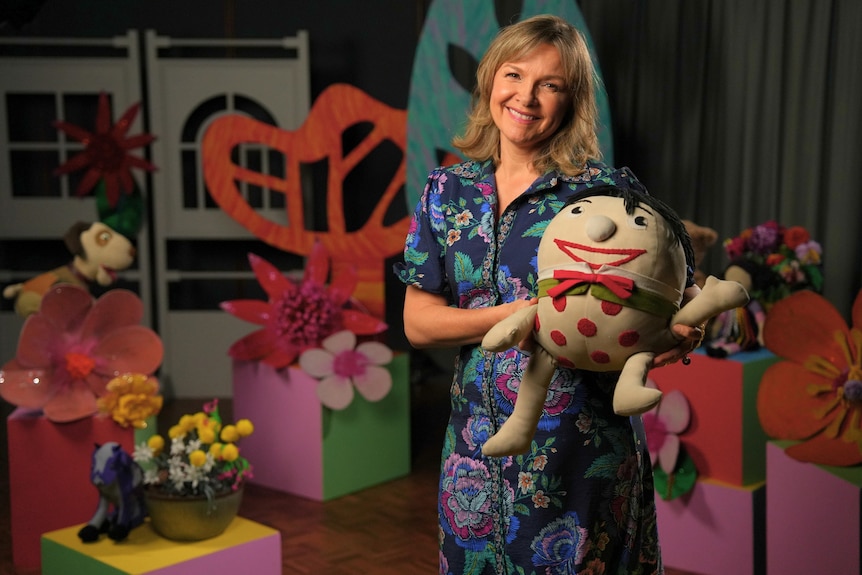 A smiling blonde woman stands on a colourful TV set of Play School holding 'Humpty' the round spotted toy