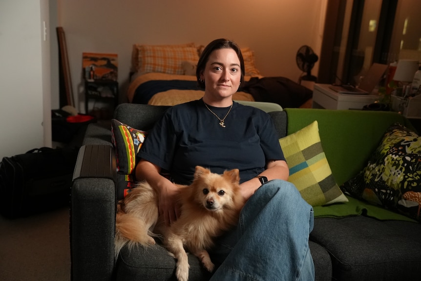 A young woman sits on a couch in a studio apartment with a small dog. Her facial expression is serious.