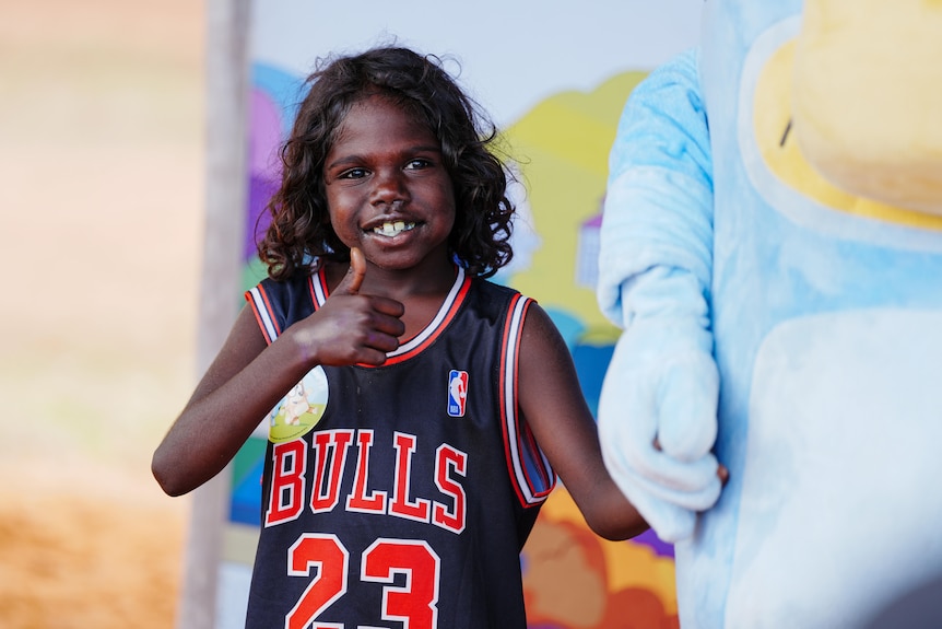A smiling boy with long, curly hair gives the thumbs up as he stands next to a person in a Bluey suit.