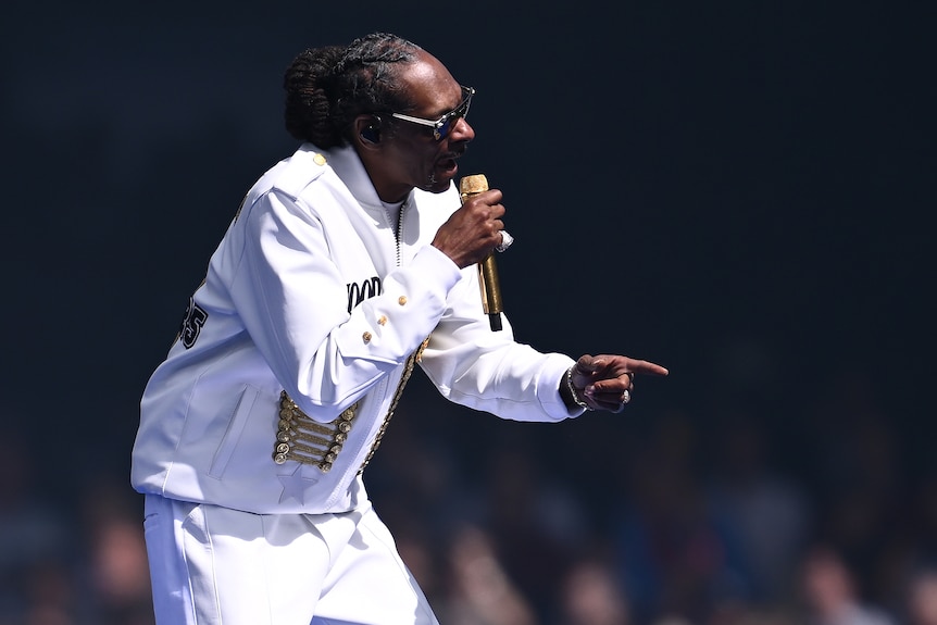 A man with his dreadlocked dark hair pulled back behind him, dressed in white in dark glasses holds a gold microphone.