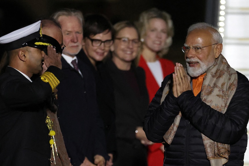 A man holds his hands together in a praying symbol as he looks at six people standing in a line.