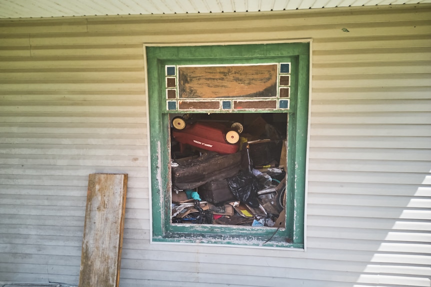 A green-framed, open window in the side of a white weatherboard home, crammed full with a pile of discarded rubbish.