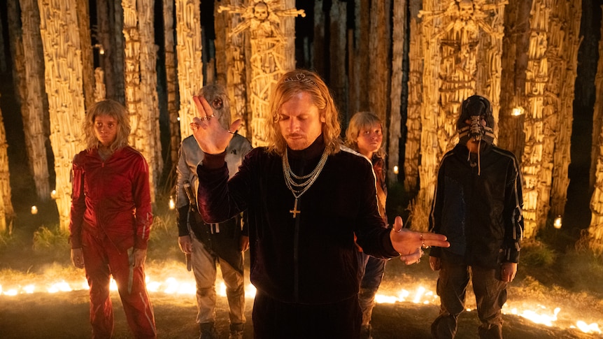 Jack O'Connell in a long blonde wig stands in a ring of fire with two hands raised in finger gun shapes.