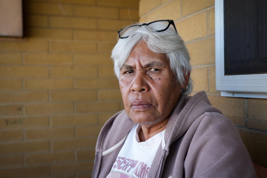 An older woman with white hair sits in the corner of a brick building.