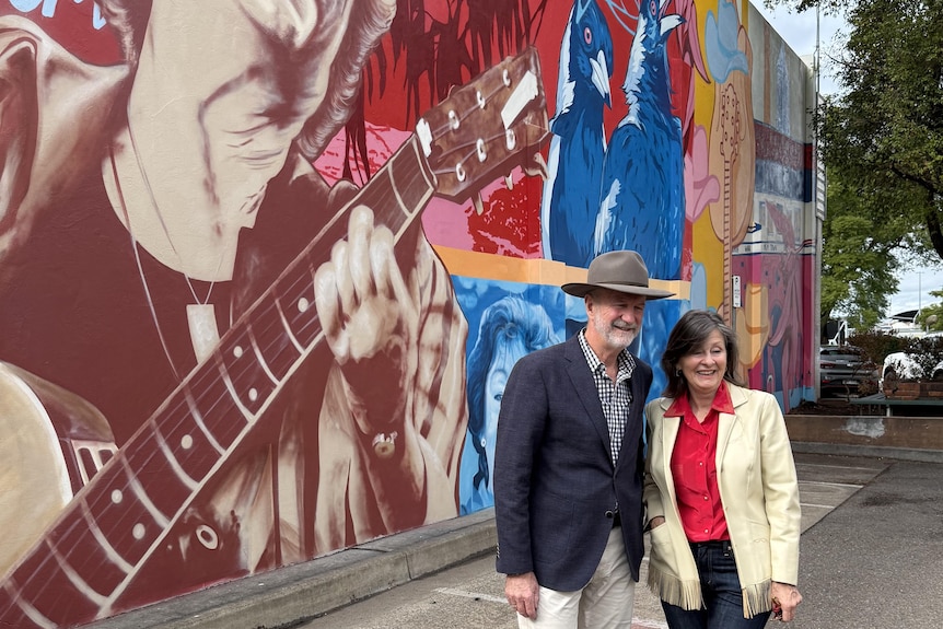 Two siblings stand in front of a mural of their mother smiling at the camera