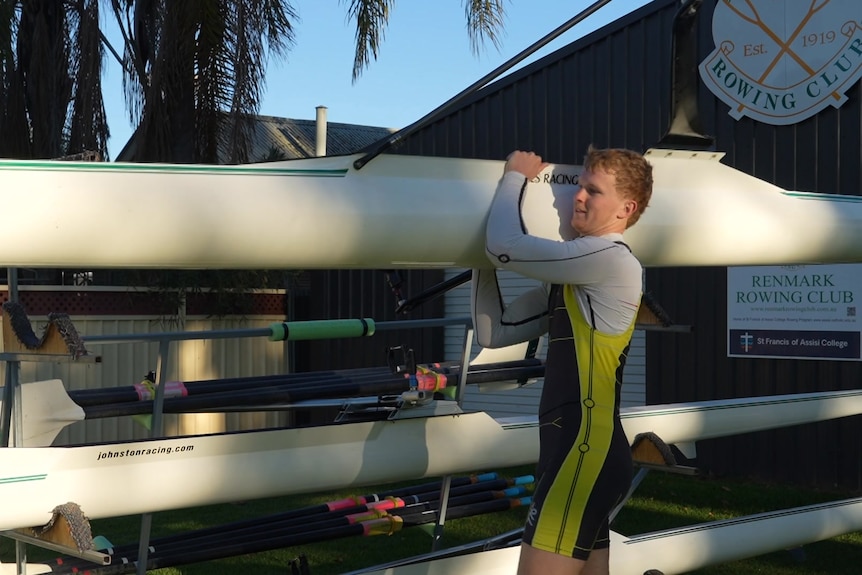 A man wearing a wetsuit carrying a white row boat.