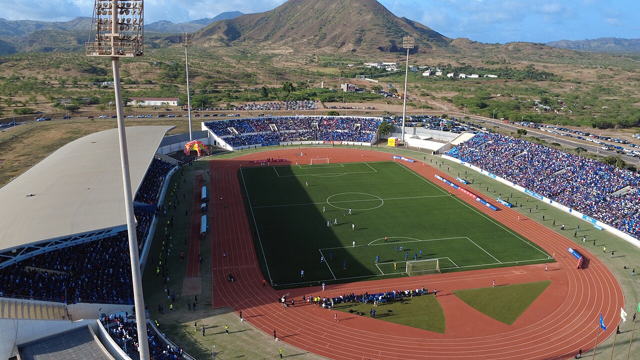 Aerial view of Cabo Verde’s Estádio Nacional filled with supporters during an international football match, with mountains rising behind the stadium as play unfolds on the pitch.