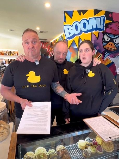 Three people stand in a restaurant with a sign behind them saying 'boom'