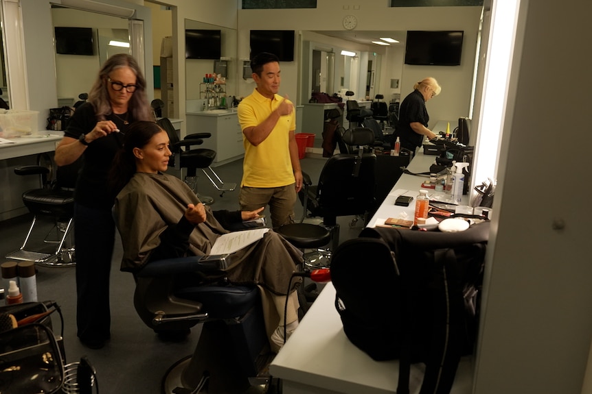 A woman sits in a hairdresser chair with cape on as woman does her hair. Man stands next to them