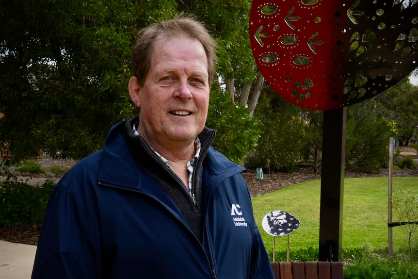 A man in a navy blue jumper with the Adelaide University badge and logo on the left breast. He is standing in a park.