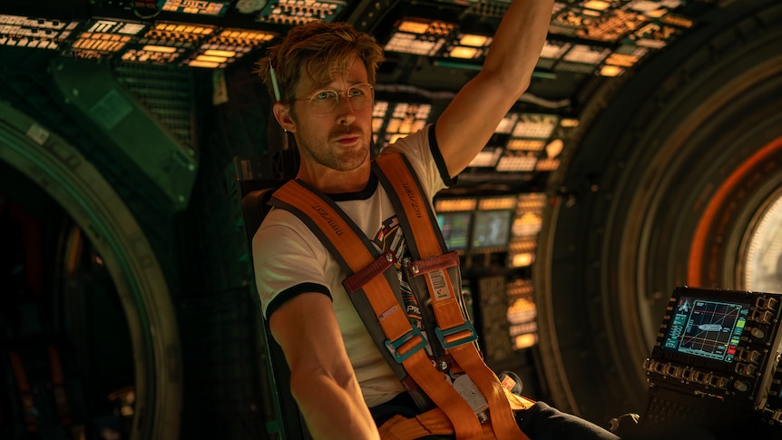 Ryan Gosling sits and braces himself in a rocket cockpit with an orange shoulders strap belt on