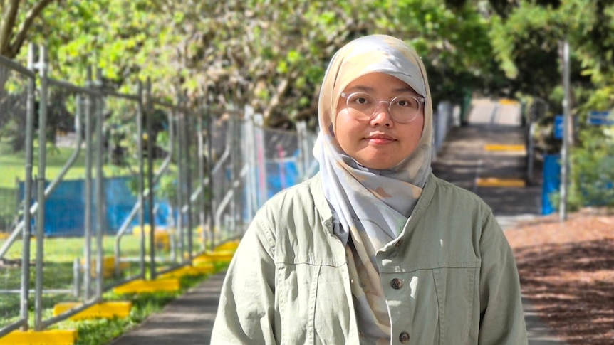 A young woman wearing a headscarf and a winter coat stands by a tall construction fence along a path.