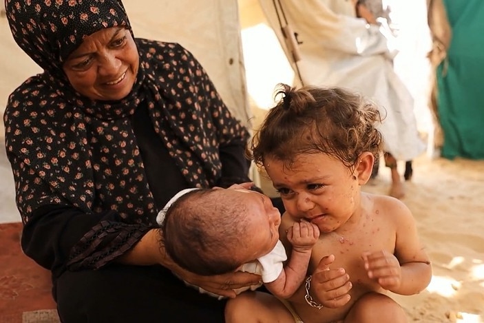 A toddler smiles while looking at her baby sister in the hands of their mother.