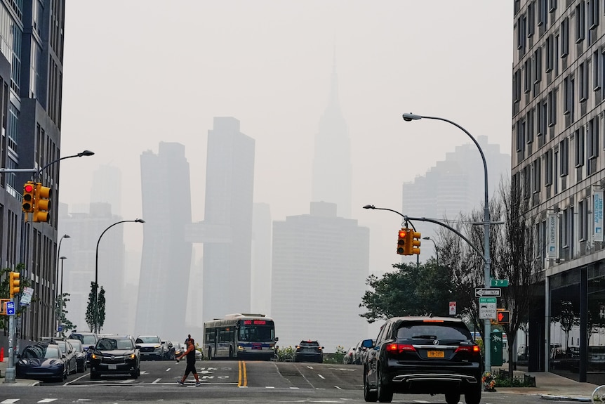 Pedestrians cross a busy street as the skyscrapers in the background are shrouded in grey haze.