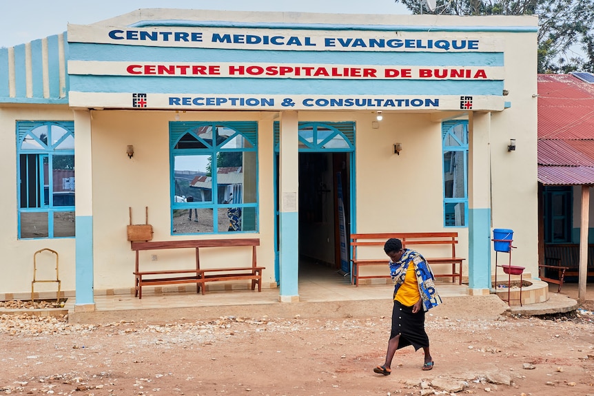 A woman walks past a pale yellow building with a sign that indicates it is a medical facility