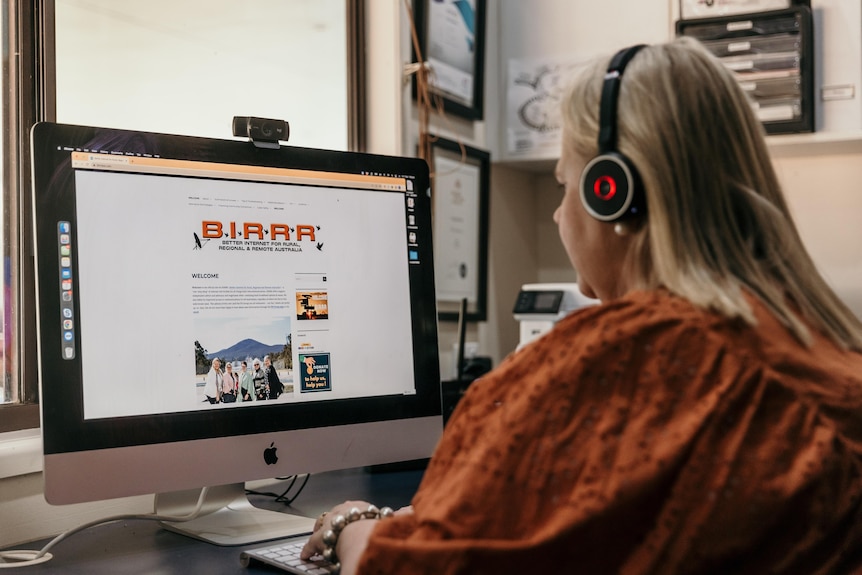 Woman sits at a desk with headphones on, facing a computer