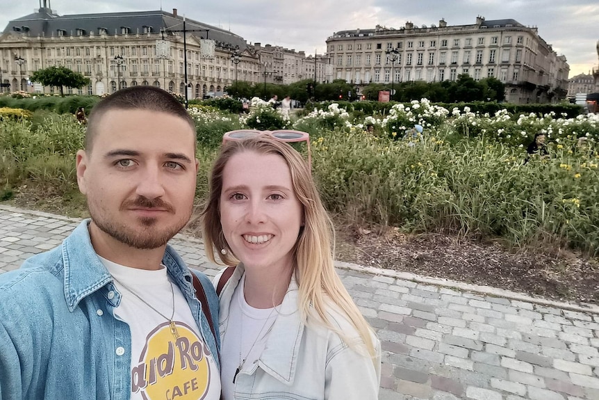 A man and woman stand together with a grand historical building in the background. She has long blonde hair.