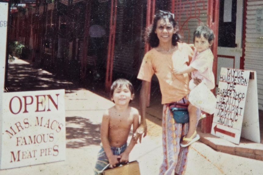A lady holding a baby with a smiling toddler next to her, placards saying Open, Mrs Macs famous meat pies.