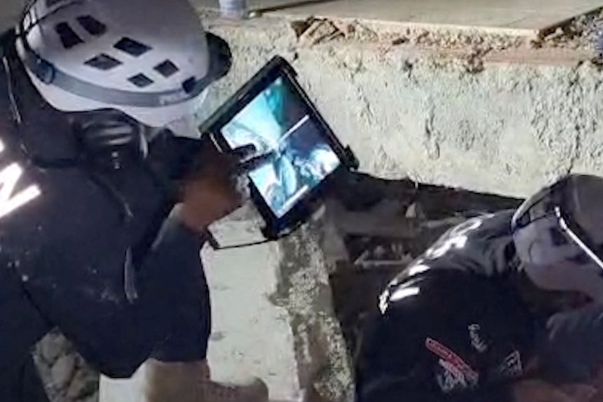 Rescue workers in dark clothing and white helmets, using a camera screen and kneeling next to collapsed rubble.