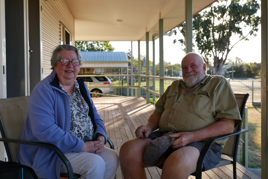 A man and a woman sit in chairs on a front porch of a house on a sunny day.