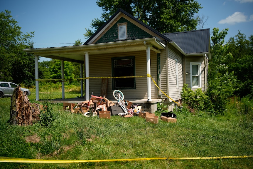 A white, weatherboard home with a grey tin roof and green lawns surrounded by yellow police tape with a pile of rubbish.