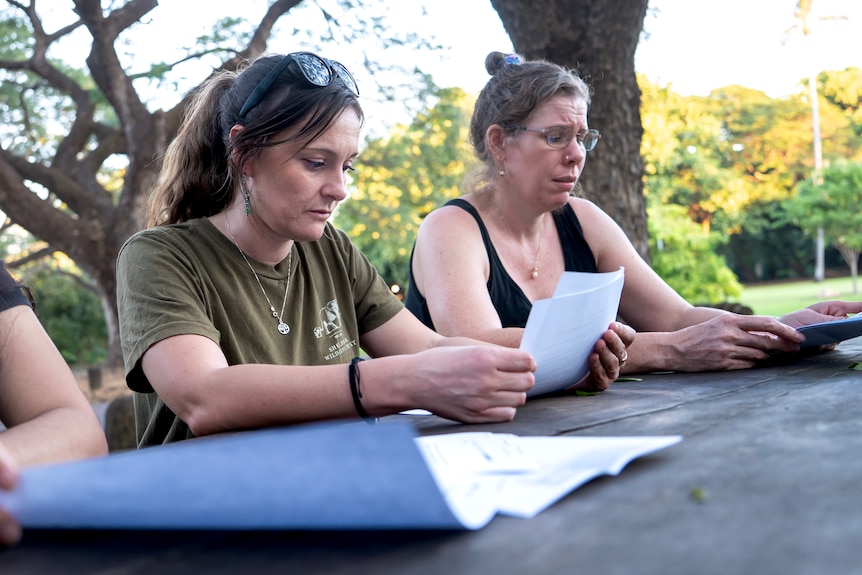 Two women sitting at a table both looking down at paperwork, with serious and upset expression.