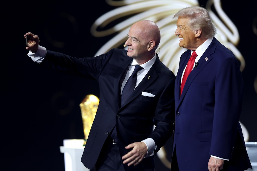 A man holds a camera up to take a selfie with a second man smiling beside.
