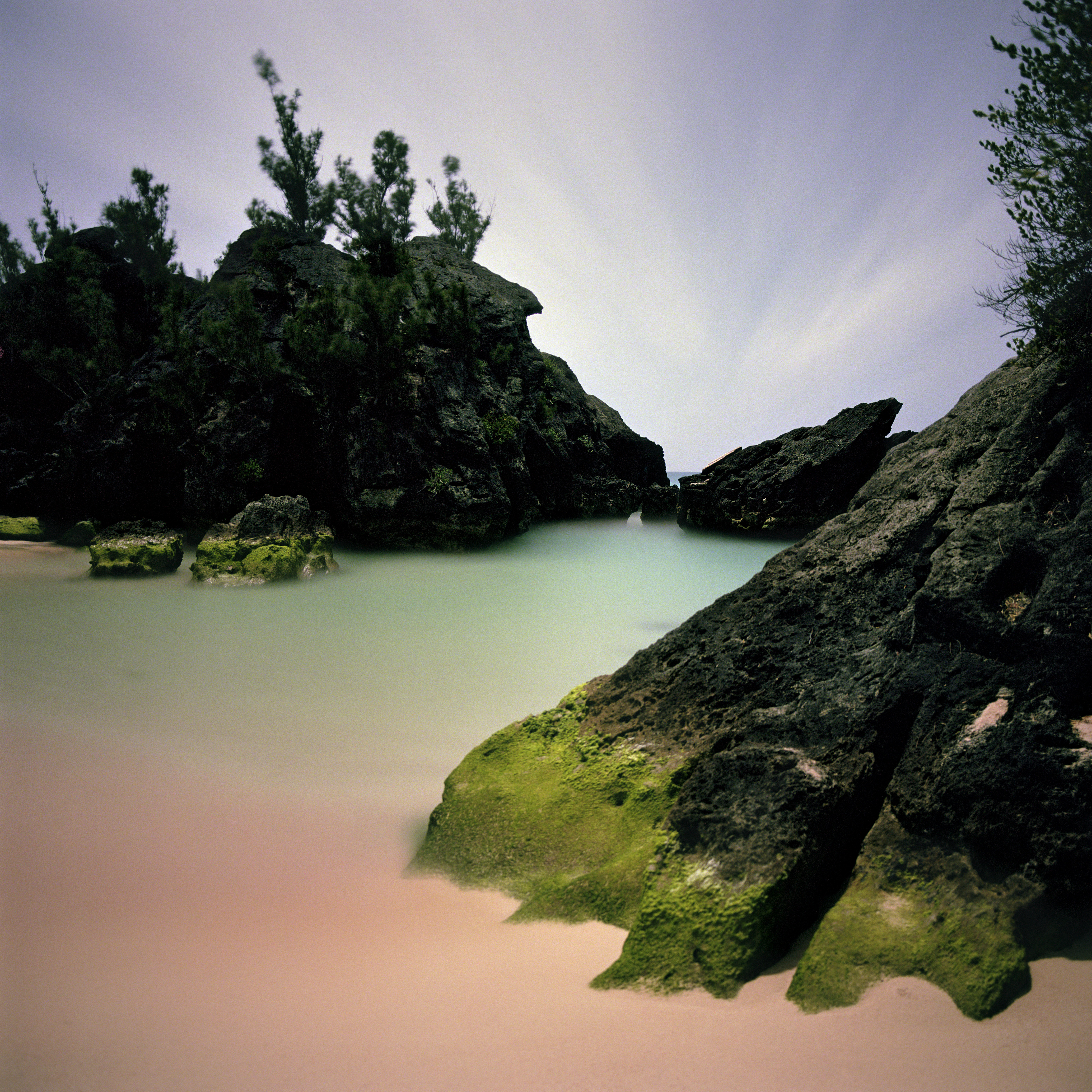 A close-up view of a secluded cove with moss-covered black rocks framing calm, pale green water and a soft pink sand beach.