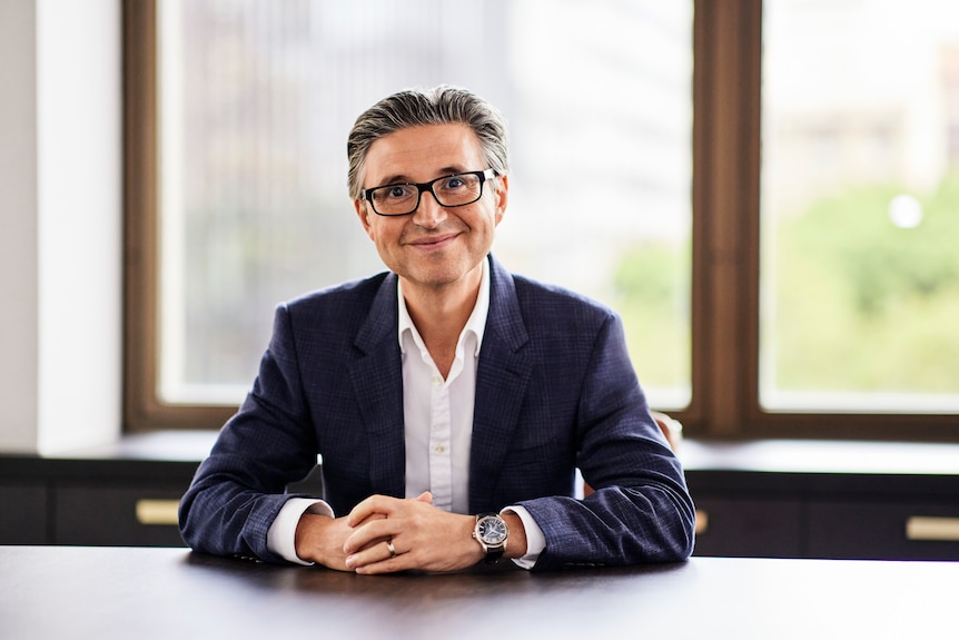 A man in a suit and glasses sits at a table with his hands resting on top of it.
