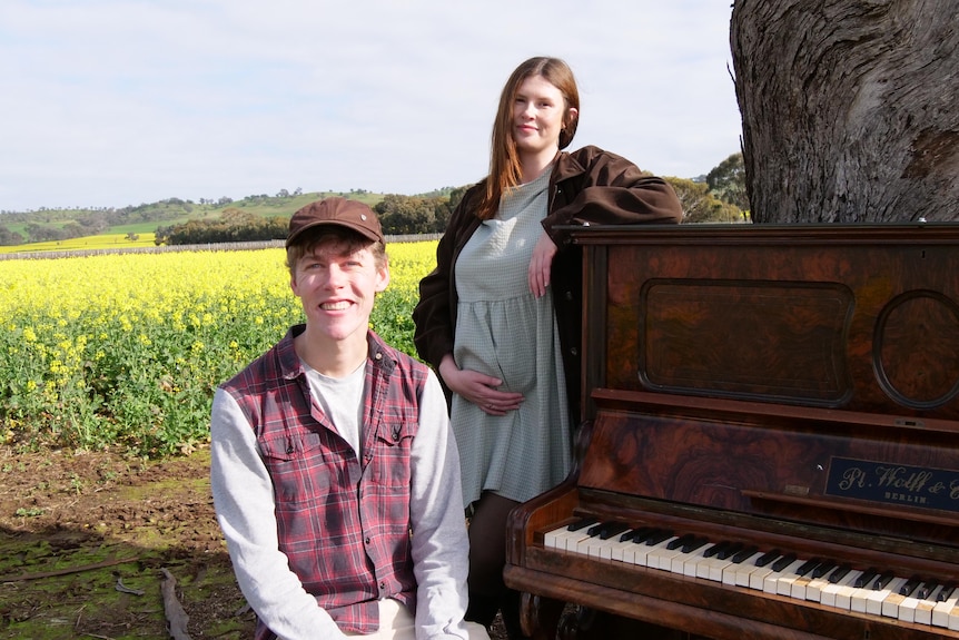 A man and a woman pose near a piano in a canola field