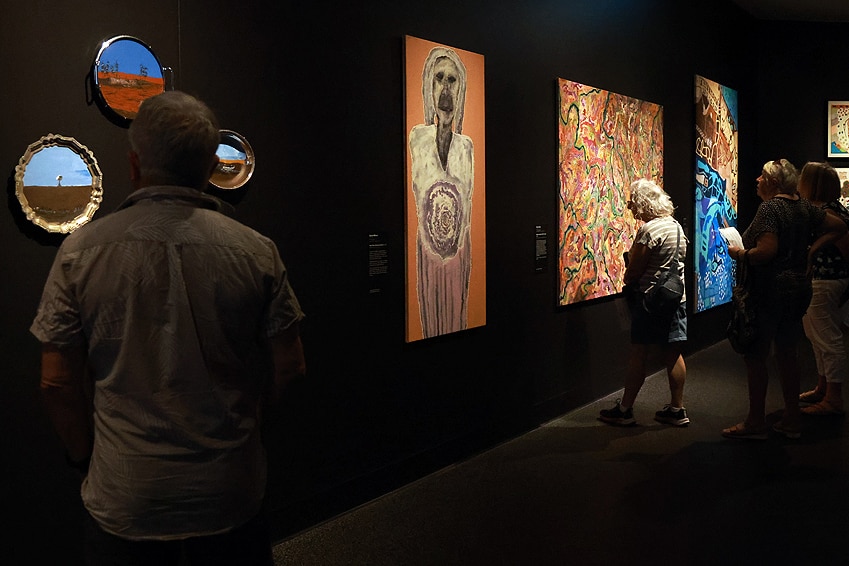 People inside an art gallery inspecting the works on display, many of the paintings feature red dirt and orange hues.