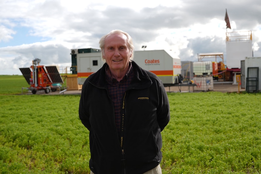Man in a dark jumper standing in green field with worksite equipment behind him.