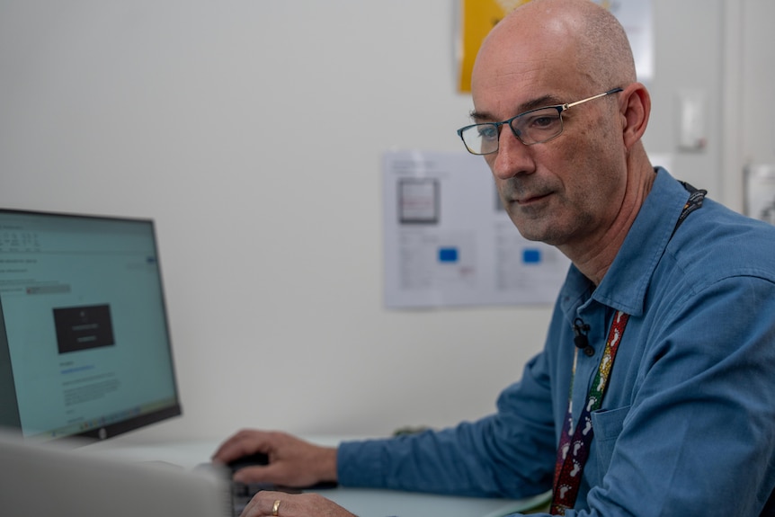 A man is bald and wears glasses and a blue shirt, he looks at documents sitting in front of a computer
