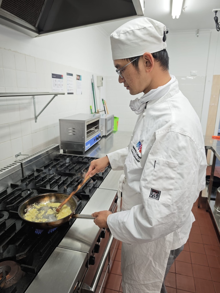 A man wearing glasses and a white chef's uniform is stir-frying at the stove.