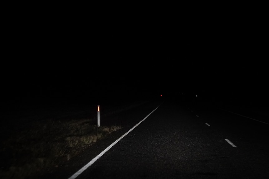 A dark lit image of the side of an outback road, with dim white light shining on the white paint lines and road marker pole.