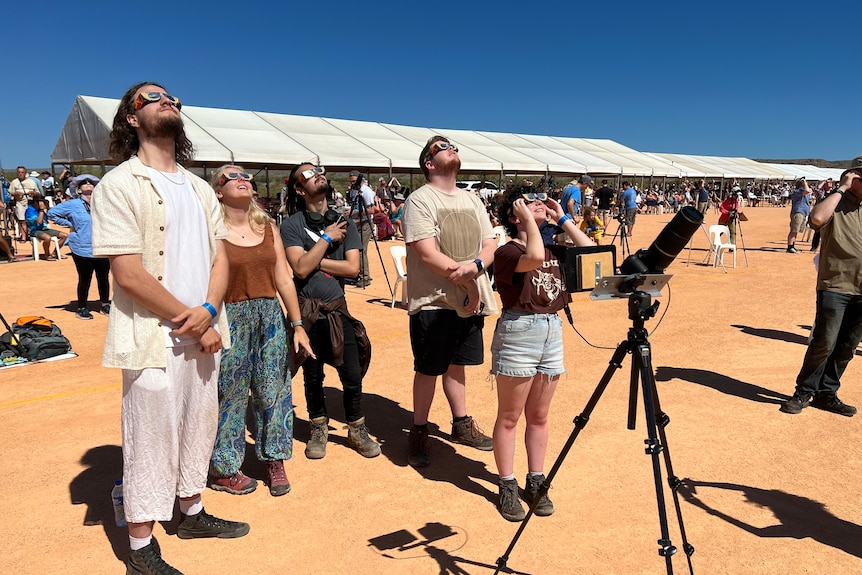 A group of people and telescopes standing in daylight looking up at sky