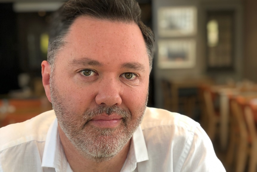 Mr Kavanagh in a white shirt smiles at the camera while sitting in a cafe or restaurant.