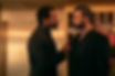 Two men in black suits face off indoors, one pointing at the other, with warm lighting and flowers in the background.