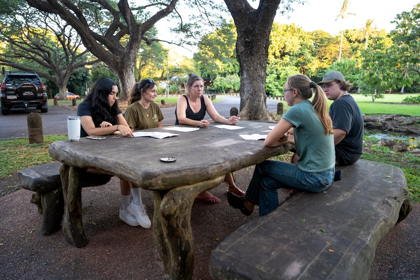 A group of five people sitting at a table bench looking at each other, in a green botanical area.