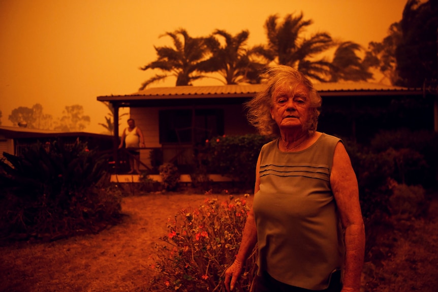An exhausted woman stands outside her home as both she and the landscape are bathed in red from nearby bushfires.