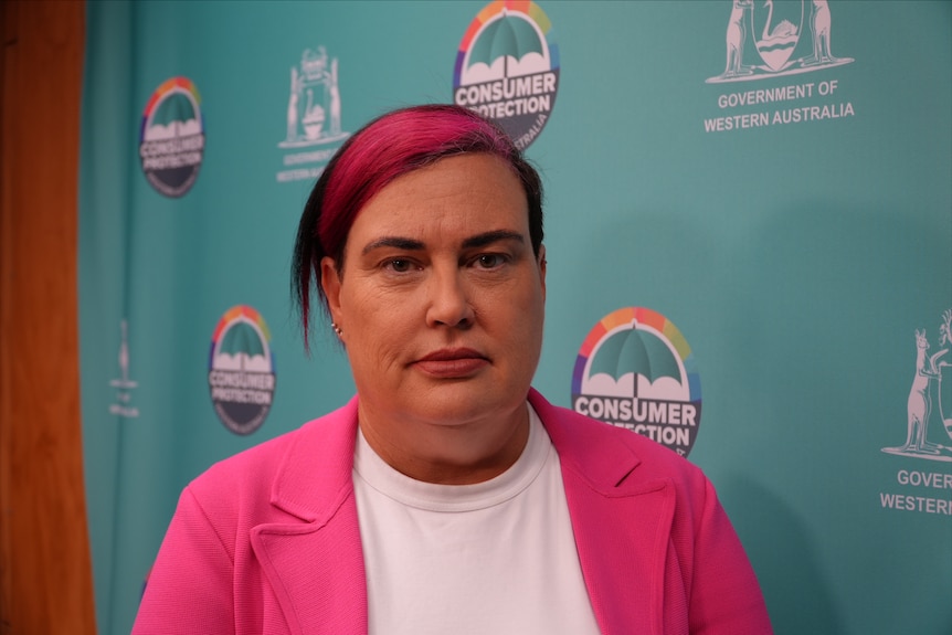 A woman with pink hair and a blazer stands in front of a sign