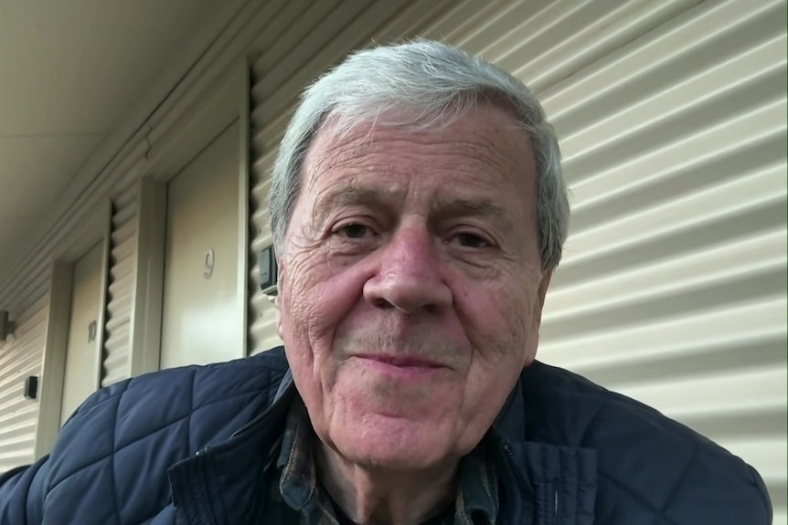 A white haired man in a navy puffer jacket stares at the camera.